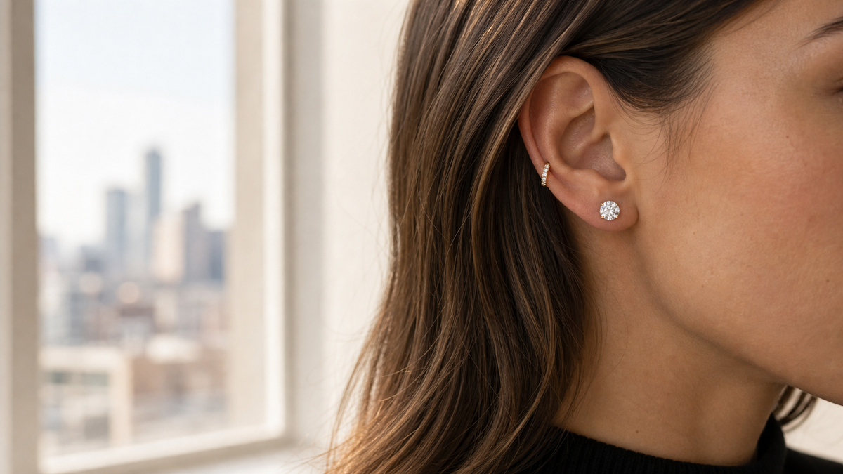 Diamond stud earrings and a small diamond huggie worn by a window in soft daylight.