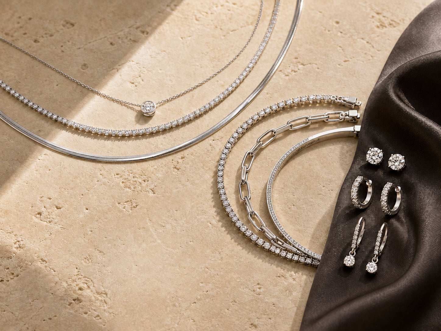 Layered diamond necklaces, bracelets, and three pairs of earrings arranged on warm ivory stone